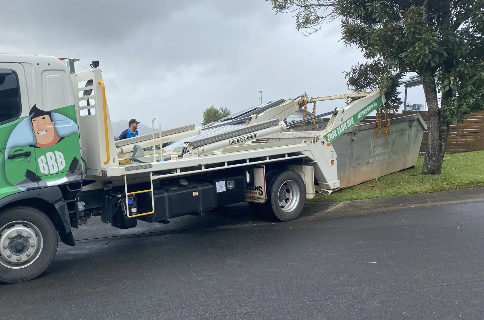 Big Boyz Binz truck setting a skip bin down on a lawn beside a tree and a fence in Cairns